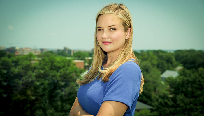 A woman in a blue top with trees in the distance stares at the camera.