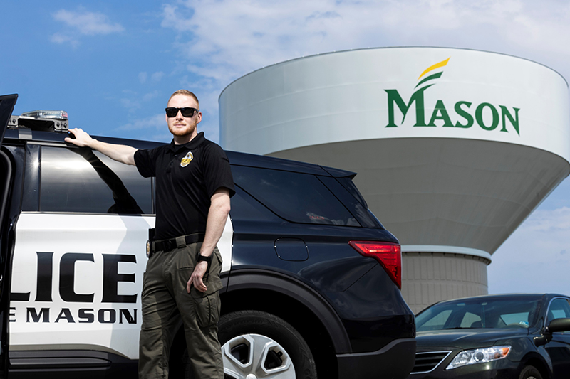 A man in a police uniform and wearing sunglasses stands next to a police car with the Mason water tower behind it.