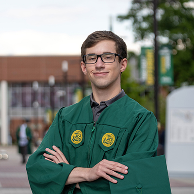 Brad stands with arms crossed in green graduation robes on Wilkins Plaza