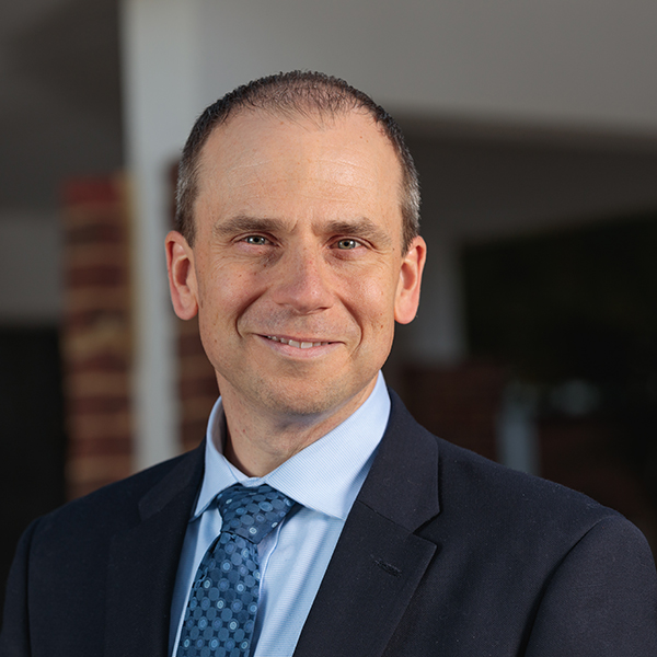 Smiling man wearing light blue shirt with navy tie and jacket.