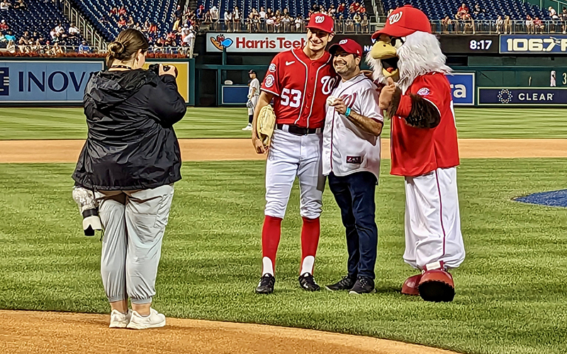 A man in a white shirt holds a baseball while a man in a red baseball uniform and an eagle in a baseball costume pose with him for a photographer.