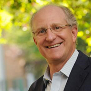 A man in glasses and a striped shirt stands in front of a tree and smiles.