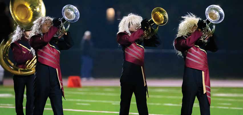 High school band members in red uniforms and 80's style heavy metal wigs perform on a football field.