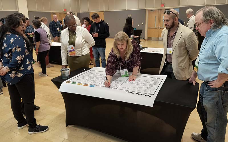 Several people watch as a woman with a nametag writes something on a large poster on a table.