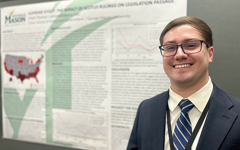 A young man wearing glasses and a navy suit jacket and tie stands in front of a research poster.