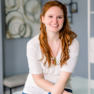 A woman with red hair in a white shirt sits and smiles