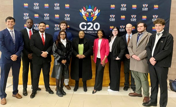 A group of well-dressed people stand in front of a sign advertising the G20 summit.