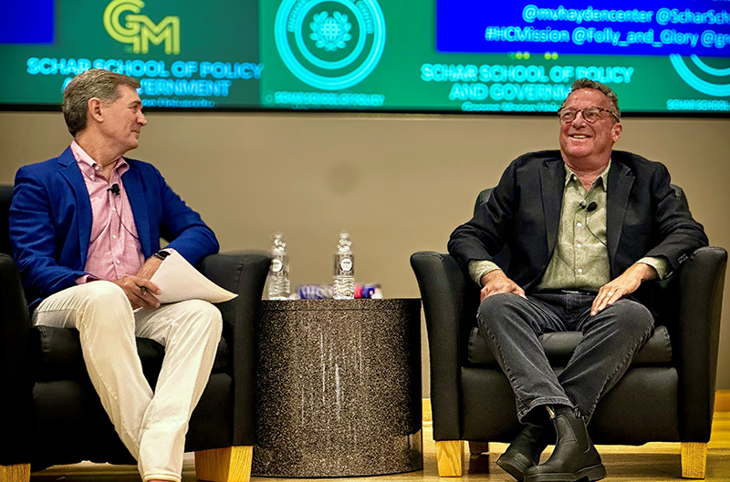 Two men sit on chairs on as stage with a green and gold background.