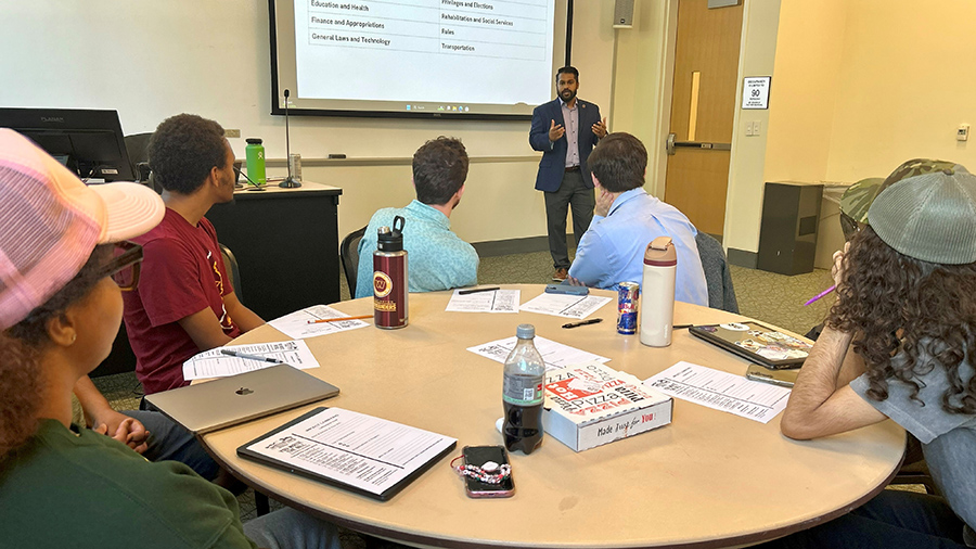 A man stands in front of a screen with a table of people sitting in the foreground.