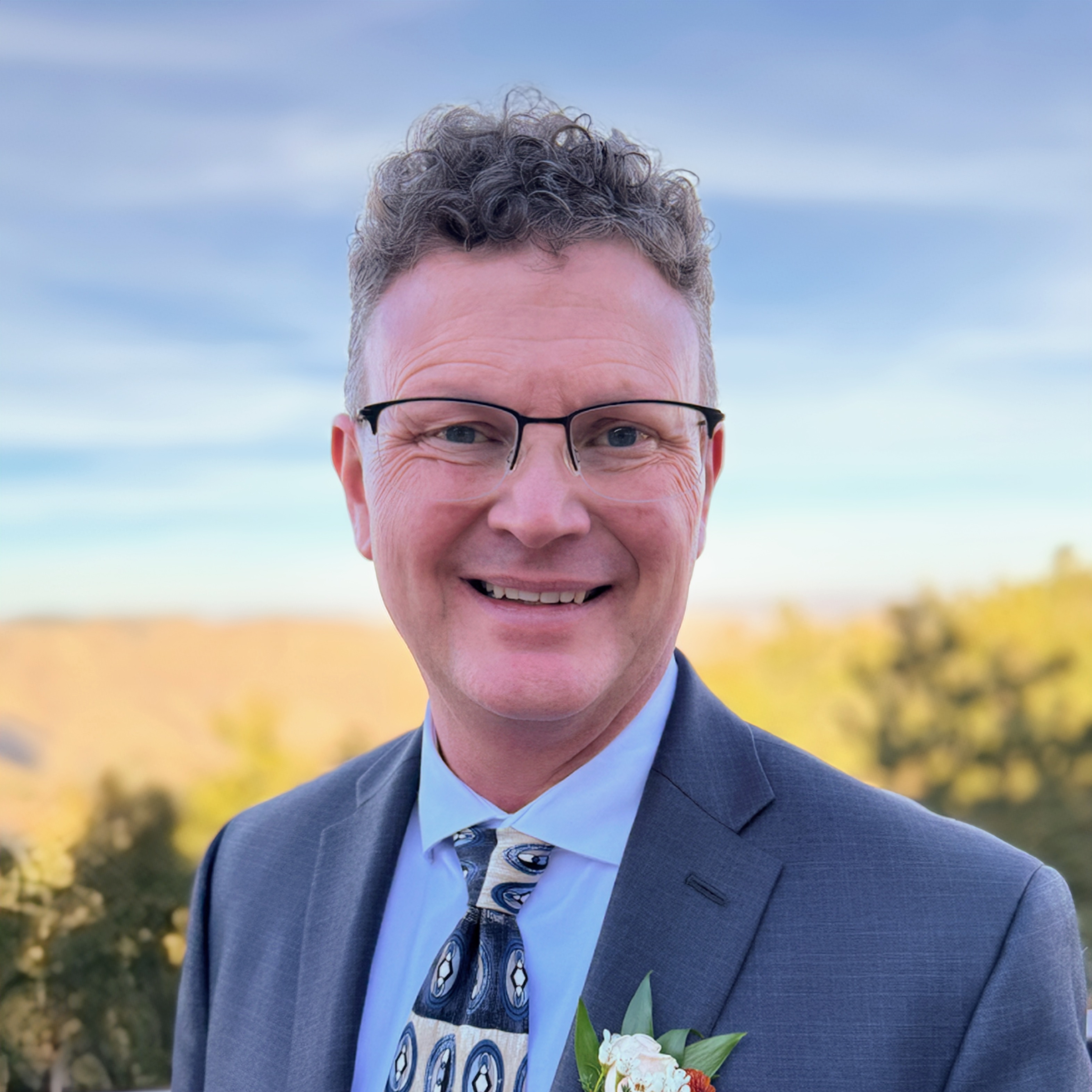 A headshot of Robert Pettit standing outside and wearing a light blue collard shirt, a suit jacket, and patterned neck tie.