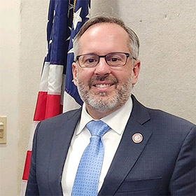 A man in a gray suit, white collared shirt, and blue tie stands in front of an American flag.