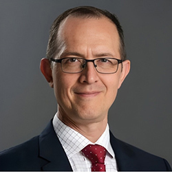 A man with glasses stands and smiles while wearing a black blazer, white collared shirt, and red tie.