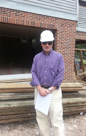 ID: A man in a white hard hat, sunglasses, and a purple button up shirt stands in front of a building with construction materials in the background.