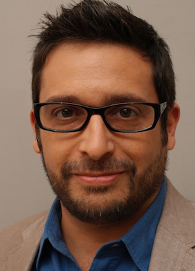 Picture of Arthur Romano, a man with tan skin, brown hair, glasses, and a brown beard. He is wearing a blue collared shirt and a tan blazer.