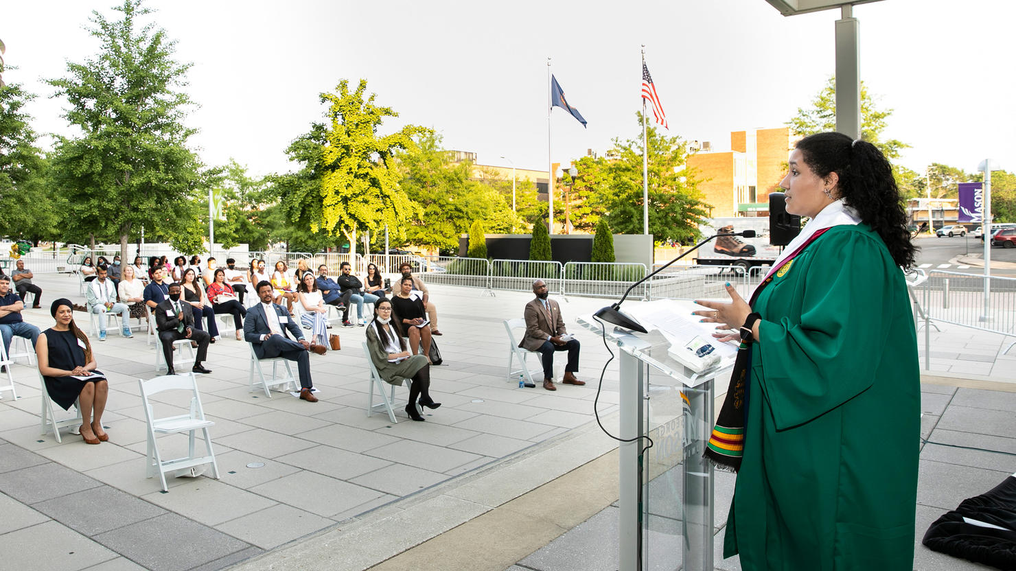 A socially distanced crowd of seated people on Mason Square's Plaza listen to a speaker in graduation robes.