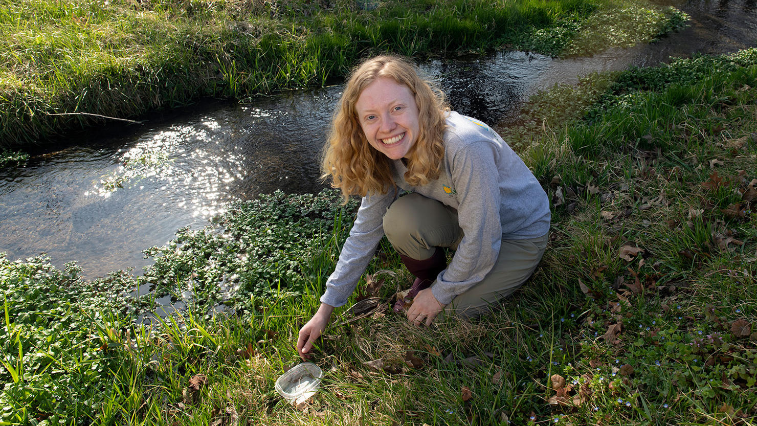 Maggie Walker sits by a stream with a sample of water in a small tupperware. 
