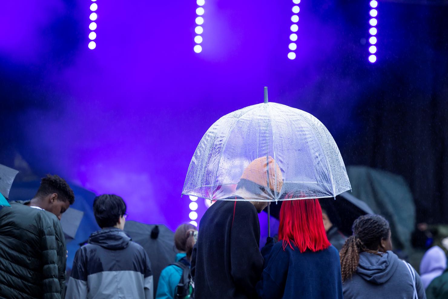 Two people stand close together under a transparent umbrella within the crowd, in front of the performance stage at Mason Day. The rain is coming down.