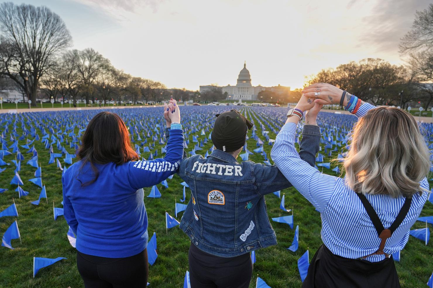 Three people stand together with their arms raised in front of thousands of blue flags on the National Mall in front of the U.S. Capitol