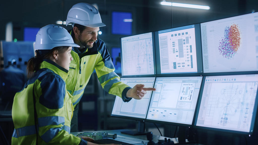 Two systems engineers, a man and woman, evaluate data on a computer screen while wearing yellow jackets and hard hats