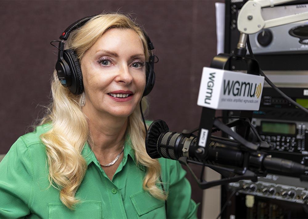 Karina Korostelina wears a green blouse and necklace. She has earphones on and looks at the camera. A microphone is just to her right.