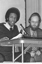 Andrew Evans at left talking into microphone with unidentified male next to him