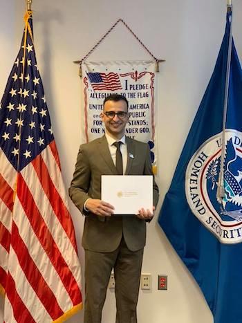 Haider Semaisim wearing a suit and tie flanked by the American flag and the Department of Homeland Security flag. He is holding a certificate to show his U.S. citizenship.