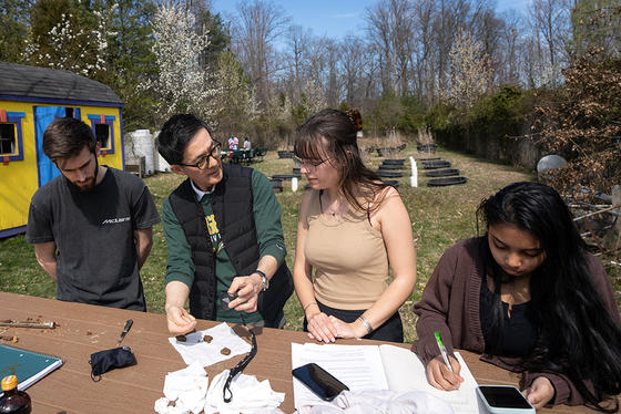 Students stand beside an outdoor table with their professor, discussing soil samples. Behind them are the plots the soil was taken from.