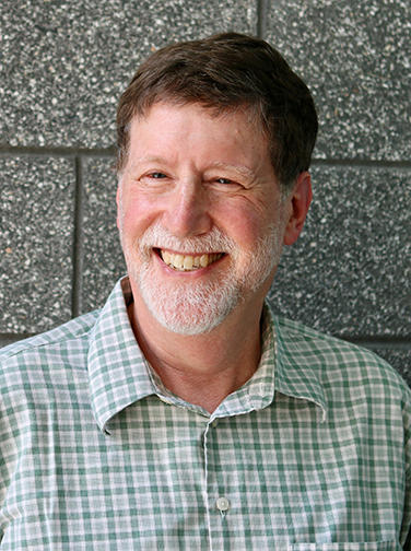 A man in a green and white checked shirt, with a short white beard and dark brown hair, smiles at the camera.
