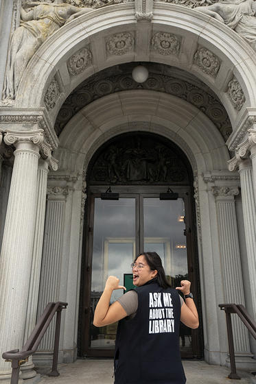 Kevynne points to the back of her vest which reads "Ask Me About the Library" while she stands on the steps of the Library of Congress