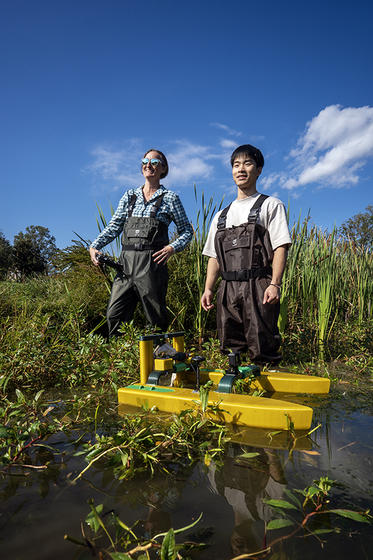 Professor Leigh McCue and student Zesen Li stand waist-deep in a pond beneath a bright blue sky. Their yellow surface water vehicle floats in front of them. 