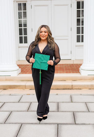 Ashley stands in a black jumpsuit with green graduation cap in her hands in front of white columns and brick walkway.