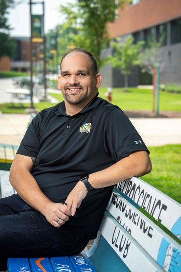 TJ Pegg sits on a bench and smiles on the quad near SUB I on the Fairfax Campus on a sunny day
