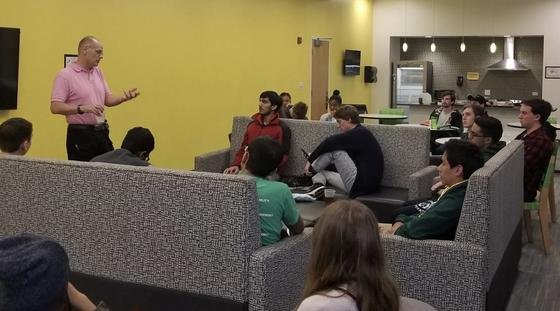 A professor addresses a group of students in a dorm common room.