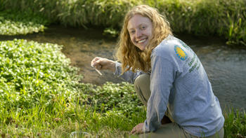Undergraduate Maggie Walker sits beside a stream of water, smiling at the camera. She is wearing an SMSC shirt and holding a QuanTab strip to measure the levels of chloride in the stream.