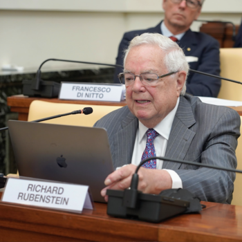 George Mason University Carter School professor Richard Rubenstein speaks at the Pontifical Institute for Social Sciences at the Vatican. He is seated at a table, wearing a gray suit and tie, and reading into a microphone. His name is shown on a desk plate. Behind him is Francesco Di Nitto.
