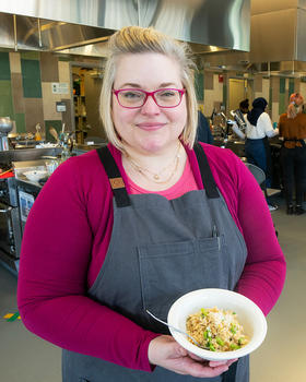 Rebecca Jones holds a bowl of risotto and smiles at the camera. Behind her, students work on their own recipes.