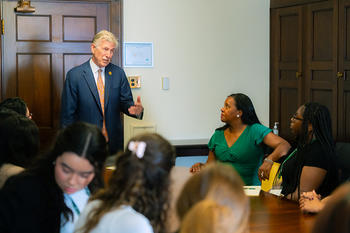 A man in a blue suit gestures as seated women listen.