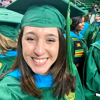 A woman in a green cap and gown smiles at the camera in a selfie.