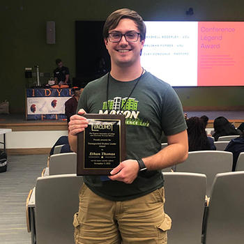 A young man in shorts and a T-shirt holds a plaque in an auditorium.