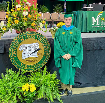 A man in a green cap and gown stands in front of a stage with flowers and an official university seal.