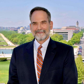 A man with a beard in a suit and tie stands in front of the Washington Monument in the distance.