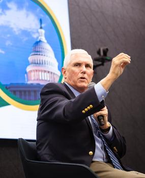 A man with white hair holds a microphone and gestures with his other hand.
